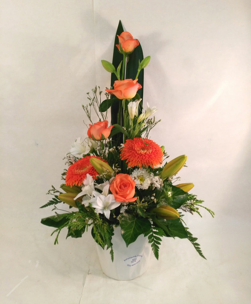 An arrangement of flowers in ceramic container, in colours of orange and white, front facing . Some roses and gerberas make up this  traditional arrangement.